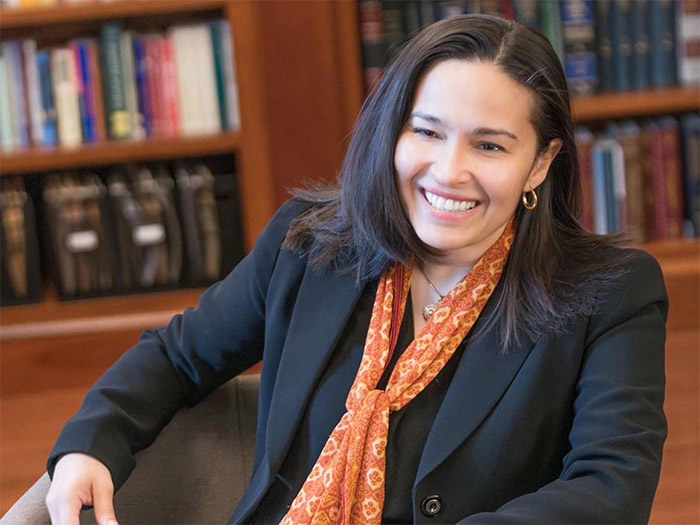 A person with long hair smiles while sitting in a chair, making strides in their black blazer and orange patterned scarf. The background showcases shelves filled with books, hinting at a journey of knowledge and growth.