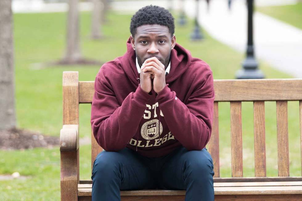 A person wearing a maroon hoodie with text sits on a wooden bench in the park, resting their hands under their chin as if waiting for a golden ticket. A walkway and trees are visible in the blurred background.