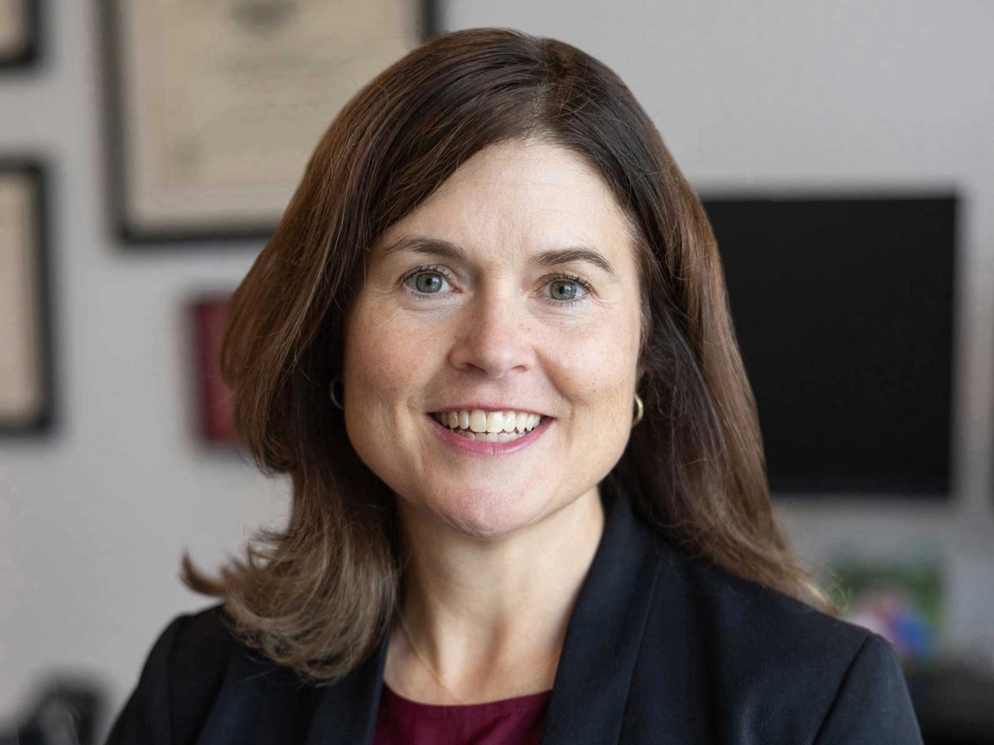 A woman with shoulder-length brown hair smiles at the camera, embodying the essence of patient care. She is wearing a dark blazer over a maroon top. The background shows blurred office items, including framed certificates, reflecting her dedication beyond the bedside.