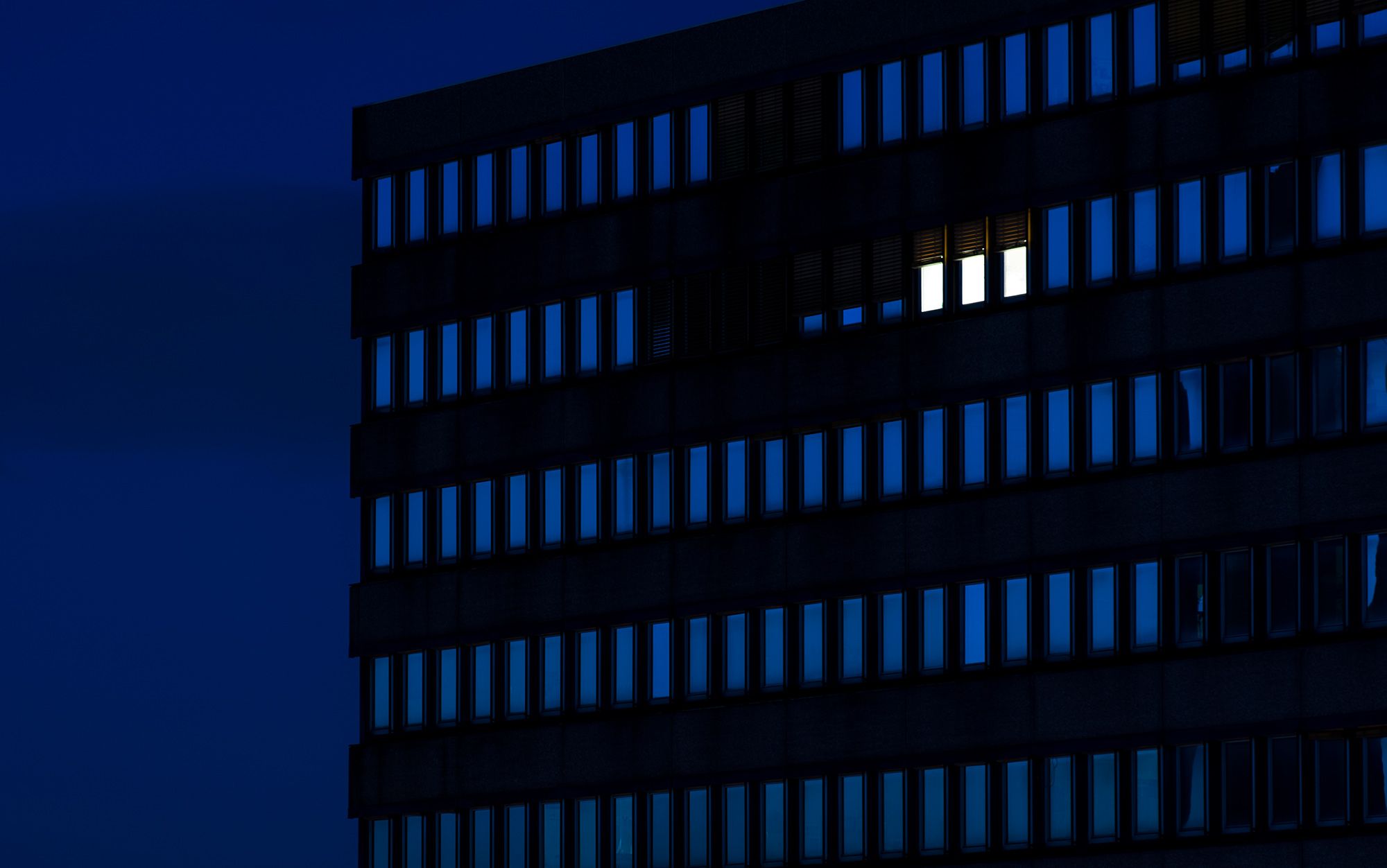 A dark building at night, the black building structure contrasting with a deep blue night sky, with many vertical rectangular windows, three windows are brightly lit.