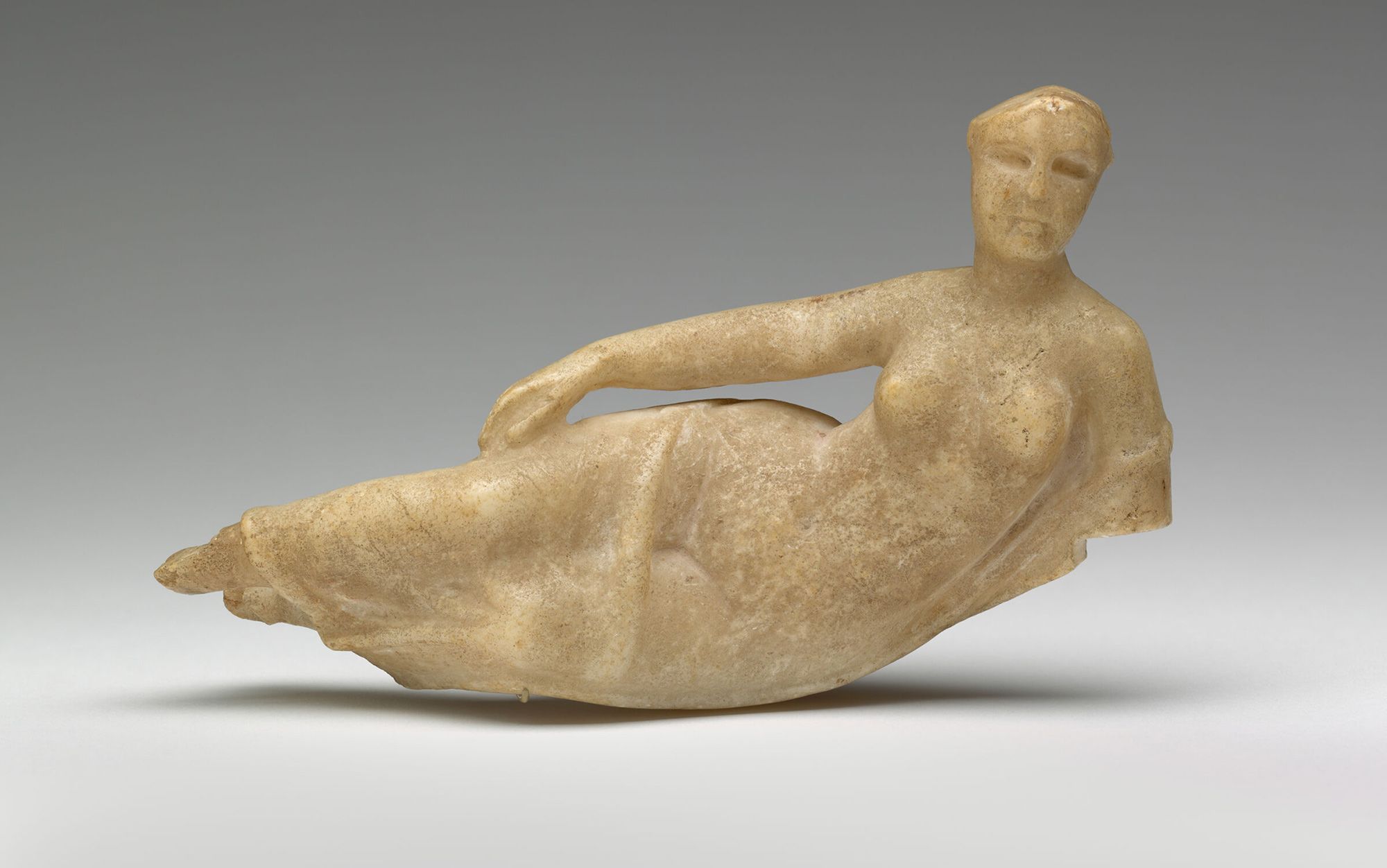 Ancient stone sculpture of a reclining female figure on a smooth surface against a grey background.