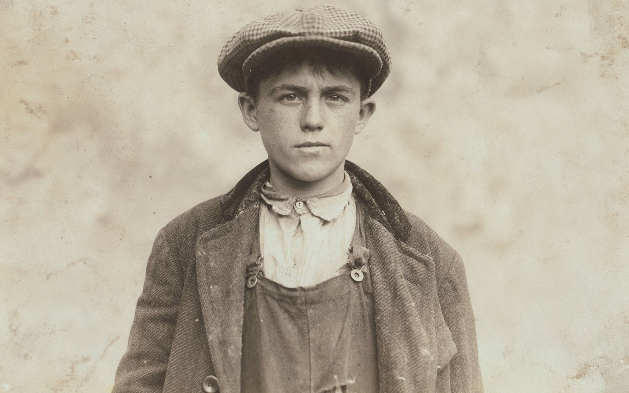 Black-and-white photo of a young boy in a cap, overalls and a jacket, standing and looking directly at the camera from a neutral background.