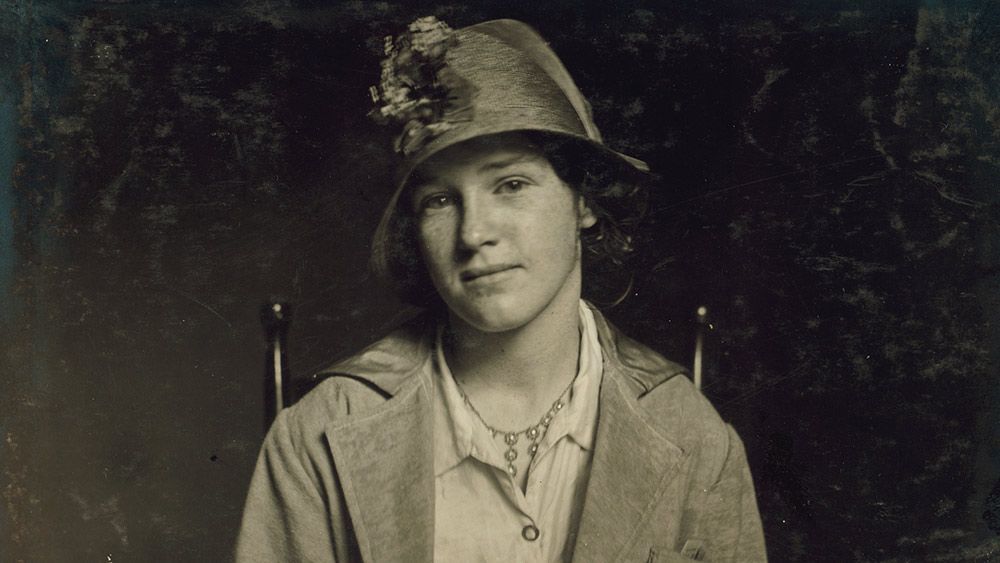 Vintage photo of a woman wearing a hat adorned with flowers, a coat and a necklace, seated and facing the camera.