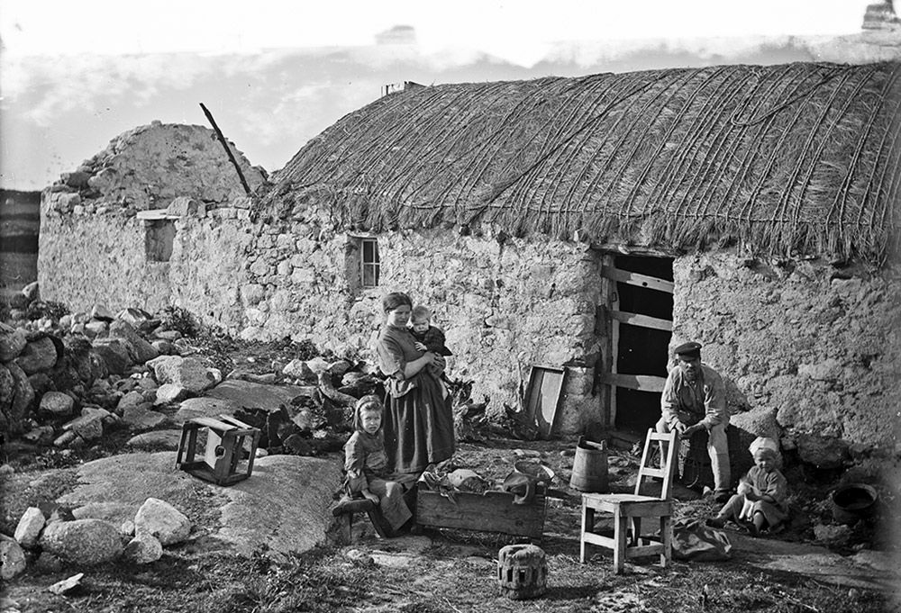 Black-and-white photo of a rural family with three children outside a stone cottage with a thatched roof among rocks and stools.