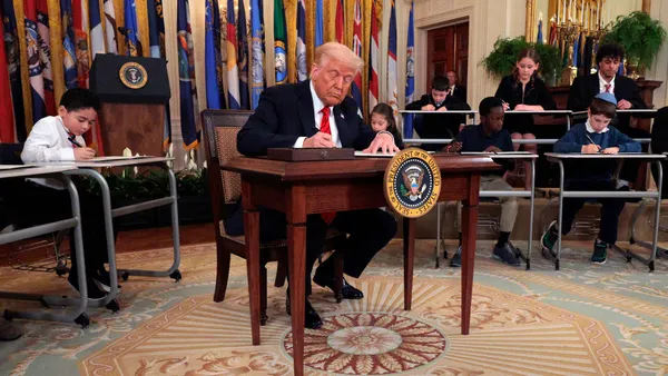 An adult sits at a small desk on a stage in a room, holding a pen and writing on a paper. Rows of children are sitting at individual desks behind the adult.