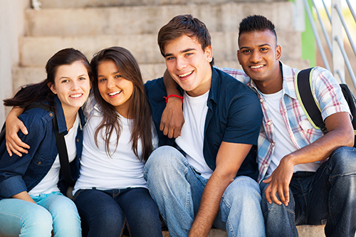 Four racially diverse teens sitting together on an outdoor bench wearing blue jeans.