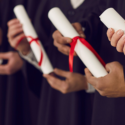 Row of graduates in black caps and gowns holding diplomas at graduation ceremony College or university students in black gowns holding traditional diploma scrolls at graduation ceremony. Happy graduates with certificates in hands celebrating milestone life event of getting degree